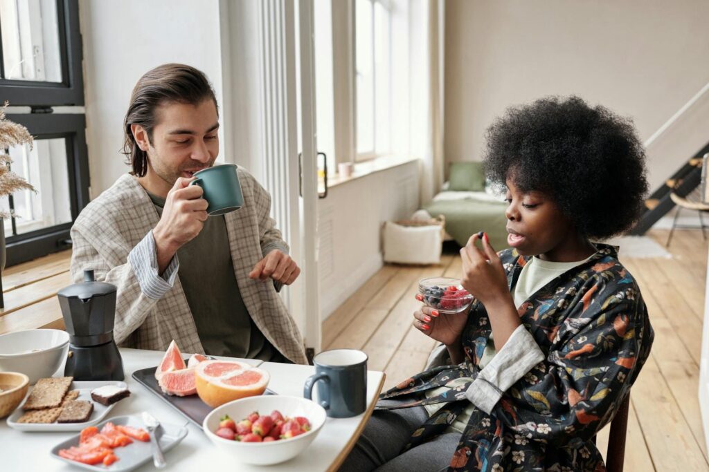 dos jóvenes tomando un buen desayuno a base de café, tostadas y fruta mientras charlan felices